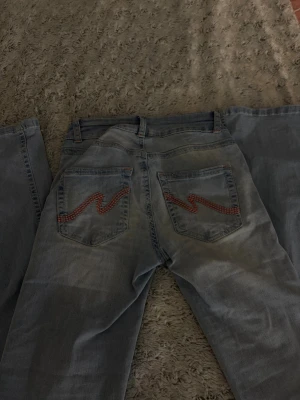 Ljusblå jeans från Nelly - Säljer ett par ljusblå jeans från Nelly med utsvängda ben och dubbla knappar i midjan. Bakfickorna har en snygg röd zigzag-detalj som sticker ut. Klassisk femficksmodell i mjukt denimtyg, perfekta till vardagslooken.