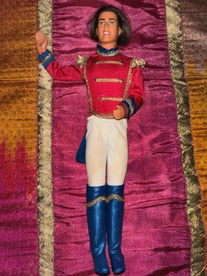 Vintage Ken prins docka – fantasy/prins-outfit – 90/00-tal - Vintage Ken-docka från sent 90-tal eller tidigt 2000-tal med klassisk prins/fantasy-look. Dockan bär röd satinjacka med gulddetaljer, vita byxor och höga blå stövlar – typisk stil från Barbie- och Ken fantasy/prins-serier från eran.  Ansiktet är fint bevarat med klar blick och välbevarad målning. Outfiten har många dekorativa detaljer och ger stark nostalgikänsla från äldre Barbie-filmer och sagoteman.  Överlag i mycket fint vintage-skick. Mindre missfärgning/slitage kan förekomma på vissa delar