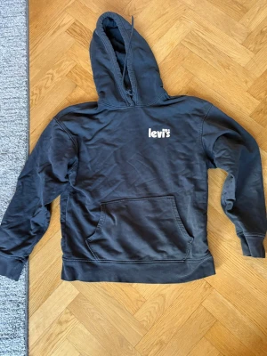 Svart hoodie från Levi's med logga - Svart/grå hoodie från Levi's med vit logga på bröstet. Klassisk känguruficka framtill och stor huva. Tillverkad i mjukt bomullsmaterial och har ribbade muddar vid ärmslut och nederkant. Något urtvättad.