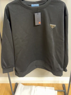 Svart hoodie från Prada - Snygg svart hoodie från Prada med klassisk rund halsringning och ikoniska metalltriangeln på bröstet. Hoodien har en clean look och ribbade muddar vid ärmar och nederkant. Perfekt för dig som gillar stilrena designer och exklusiva märken.