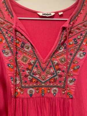 Fin! Rosa broderad klänning från Indiska - Jätte fin! Unik rosa klänning/tunika från Indiska med färgglada broderier och pärldetaljer vid halsringningen. Långärmad modell i mjukt bomullstyg med v-ringad hals och dekorativa knytband. Perfekt för dig som gillar boho-vibbar och detaljer. Härligt material i krinklad bomull. Mycket sparsamt använd i fint skick. En liten tråd på ena axeln är lite lös (inte iögonfallande men vill visa på bild) stl S men rymlig i stl så skulle säga S-M tajtast över bysten. 