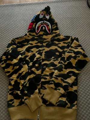 BAPE camo hoodie - Säljer en ikonisk BAPE hoodie i beige och grön camo med hajmotiv på huvan. Dragkedja framtill, ribbade muddar och detaljer i rött, vitt och blått på huvan. Perfekt för dig som vill sticka ut med streetwear och gillar unika prints.
