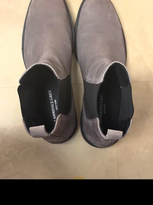 Gråa Chelsea boots från Lawrence Grey - Snygga grå Chelsea boots från Lawrence Grey i mocka med svarta elastiska paneler på sidorna och svart sula. Klassisk siluett som är enkel att matcha med olika outfits. Perfekta för dig som gillar stilrena och tidlösa skor.