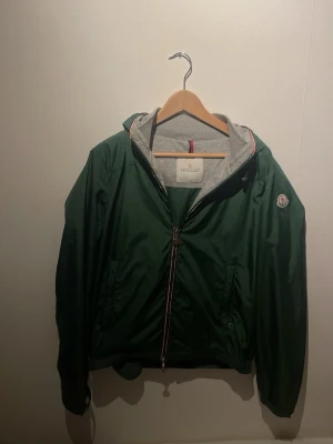 Grön vindjacka från Moncler - Snygg grön vindjacka från Moncler med huva och dragkedja framtill. Jackan har grått innerfoder, Moncler-logga på ärmen och diskreta fickor med knappar. Perfekt för dig som vill ha en stilren och sportig look.