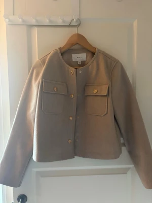 Beige cardiganjacka från Vila - Stilren beige cardiganjacka från Vila med rund halsringning och guldknappar framtill. Två bröstfickor med lock och knappdetaljer ger en trendig look. Jackan är kortare i modellen och har långa ärmar. Perfekt för lager på lager och enkel att matcha.