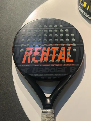 Svart padelracket från Babolat - Padelracket från Babolat i svart med stora orange bokstäver 'RENTAL' på träffytan. Racketen har rund form och många hål för bättre kontroll. Handtaget är lindat med vitt greppband. Perfekt för dig som vill spela padel med stil.