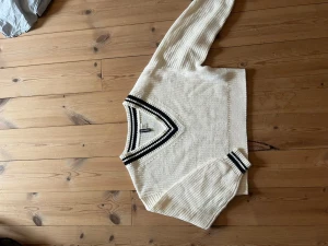 Vit stickad tröja med V-ringning Divided - Vit stickad tröja från Divided med klassisk V-ringning och svarta ränder runt halsen och ärmsluten. Tröjan har en avslappnad passform och ribbade muddar. Perfekt för lager på lager och en sportig look.