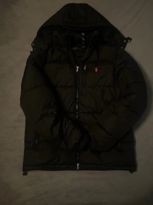 Svart pufferjacka från Polo Ralph Lauren - Snygg svart pufferjacka från Polo Ralph Lauren med huva och dragkedja framtill. Jackan har två sidofickor med dragkedja och den klassiska röda loggan broderad på bröstet. Perfekt för kalla dagar och riktigt trendig look.