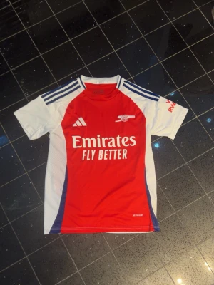 Arsenal matchtröja Adidas röd/vit - Snygg Arsenal matchtröja från Adidas i rött och vitt med blå detaljer. Tröjan har korta ärmar, klubbmärke och Adidas-logga på bröstet samt 'Emirates Fly Better' tryckt framtill. Tillverkad i lätt och ventilerande funktionsmaterial.