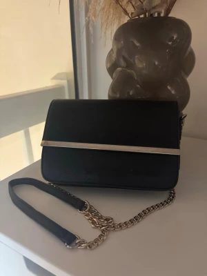 Svart axelremsväska med kedja - Snygg svart handväska med guldfärgad kedjerem och metallist på framsidan. Väskan har en stilren, rektangulär form och stängs med lock. Perfekt för dig som gillar minimalistisk design och vill ha en väska som sticker ut.