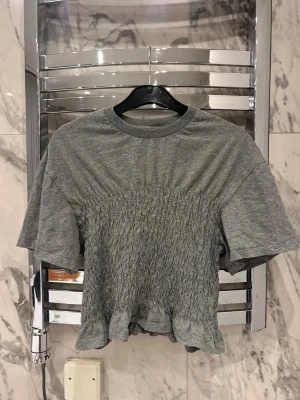 Grå croppad topp  - Säljer en grå croppad topp med korta ärmar och rund hals. Toppen har snygga rynkdetaljer över magen och en liten volangkant nedtill. Perfekt för dig som gillar en chill men ändå trendig stil.