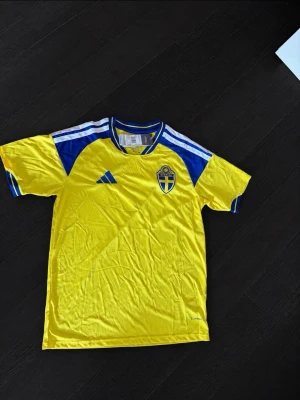 Svensk landslagströja från Adidas - Säljer en gul och blå svensk landslagströja från Adidas med klassiska vita ränder på axlarna och broderad landslagslogga på bröstet. Tröjan har rund hals och är tillverkad i lätt och ventilerande material, perfekt för fotboll eller träning.