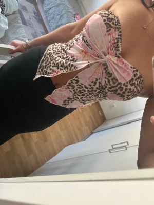 Leopard bandeau topp med rosett - Trendig bandeau topp med leopardmönster och stor rosett framtill i rosa och beige toner. Toppen är axelbandslös och har en snygg, croppad passform som ger en edgy vibe. Perfekt för dig som vill sticka ut med en unik och djärv look.