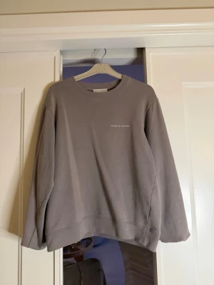 Grå sweatshirt från Tiger of Sweden - Snygg grå/beige sweatshirt från Tiger of Sweden med rund halsringning och diskret logga på bröstet. Tröjan har en avslappnad passform och är tillverkad i mjukt bomullsmaterial. Perfekt för en stilren och bekväm look.