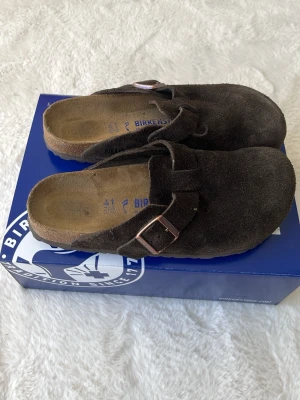  Birkenstock tofflor - Säljer ett par klassiska Birkenstock Boston tofflor i mörkbrunt mocka med justerbart spänne i metall. Tofflorna har en bekväm fotbädd i kork och mocka, perfekt för dig som gillar stilrena och tidlösa skor. Passar både hemma och ute.
