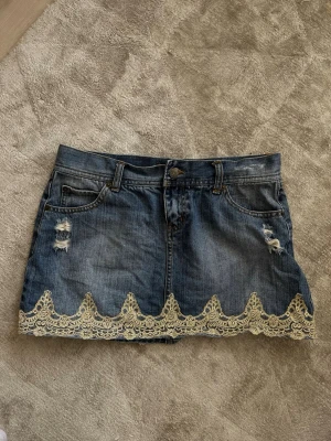 Lågmidjad Jeanskjol med spetsdetalj - Snygg kort jeanskjol i blå denim med slitna detaljer och dekorativ beige spets längst ner. Kjolen har klassiska fickor fram och bak samt bälteshällor. Perfekt för dig som vill ha en trendig och unik look. Står att det är en 38 men passar mer S/XS