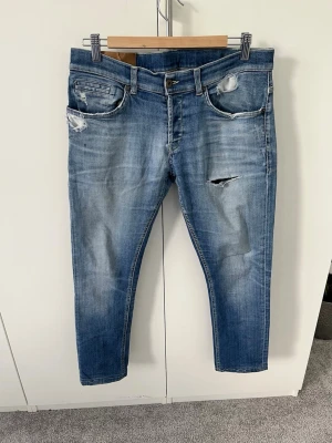 Blå slitna jeans med hål från DonDup - Snygga blå jeans från DunDop med slitningar och hål på låret och vid fickorna. Jeansen har fem fickor, smal passform och klassisk knappgylf. Perfekta för en avslappnad och trendig look.