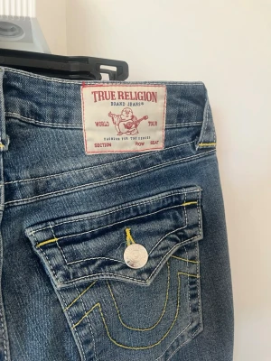 True Religion Becca Bootcut Jeans - Storlek: L31 W24/25 osäker. Snygga blå bootcut jeans från True Religion, modell Becca Mid Rise Boot. Jeansen har klassiska kontrastsömmar i gult, silverfärgade knappar och detaljerade bakfickor med logga. Perfekta för dig som gillar en avslappnad men trendig look.