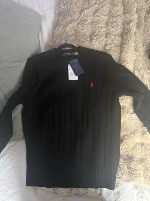 Svart kabelstickad tröja Polo Ralph Lauren - Svart kabelstickad tröja från Polo Ralph Lauren med rund halsringning och röd broderad logga på bröstet. Tröjan har ribbade muddar vid ärmslut och nederkant. Perfekt för lager-på-lager och snygg till jeans. Helt ny!