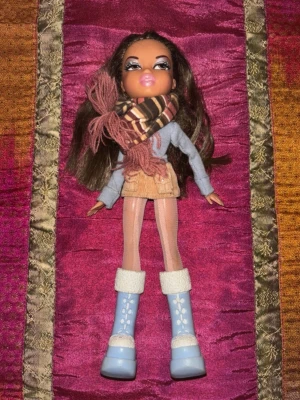 Vintage Bratz-docka – tidigt 2000-tal Y2K – original vinteroutfit - Original Bratz-docka från tidigt 2000-tal med klassisk Y2K-styling och originalinspirerad vinterlook. Dockan bär ljusblå boots med fluffdetaljer, scarf, topp och kjol i typisk Bratz-stil.  Dockan har långt brunt hår med slingor och välbevarat ansikte med den ikoniska Bratz-makeuppen. Ett fint exemplar för samlare eller för dig som gillar nostalgiska Y2K-dockor och mode från den tidiga 2000-talseran.  Mycket bra vintage-skick med fina kläder och accessoarer enligt bilderna.  Skick: Mycket bra