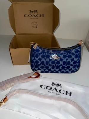 Blå monogram handväska från Coach - Snygg blå handväska från Coach med klassiskt monogrammönster i tyg och detaljer i guld. Väskan har dragkedja upptill och en avtagbar axelrem. Perfekt för dig som gillar trendiga accessoarer med lyxig känsla.
