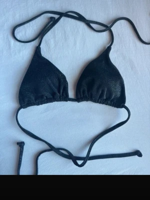 Svart glittrig trekantsbikini - Snygg svart trekantsbikini med glittrigt tyg och knytband i både nacke och rygg. Perfekt för stranden eller poolen när du vill sticka ut lite extra. Enkel och stilren design som ger en trendig look.