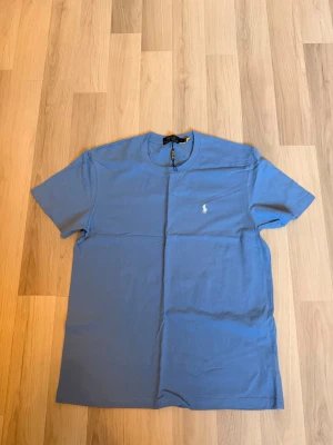Blå t-shirt från Polo Ralph Lauren - Snygg blå t-shirt från Polo Ralph Lauren med klassisk passform och det ikoniska vita broderade logotypen på bröstet. Tillverkad i mjuk bomull och har rund halsringning samt korta ärmar. Perfekt till jeans eller shorts för en clean look.