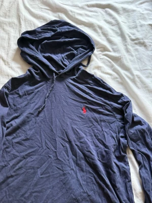 Mörkblå hoodie från Ralph Lauren - Snygg mörkblå hoodie från Ralph Lauren med klassisk röd broderad logga på bröstet. Hoodien har dragsko i huvan och är tillverkad i mjukt bomullstyg. Perfekt för en chill och stilren look.