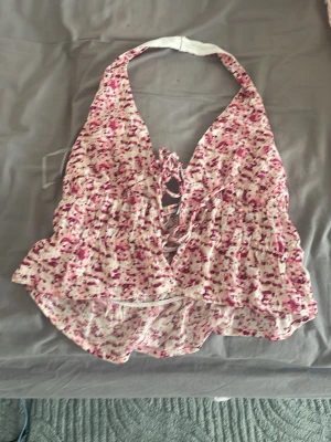 Blommig halterneck topp med snörning - Supersöt halterneck topp med blommigt mönster i rosa och vitt. Toppen har snörning framtill och volangkant nedtill. Perfekt för sommardagar och har en croppad passform. Materialet känns lätt och luftigt. Med openback 