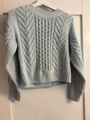 Ljusblå kabelstickad tröja - Säljer en ljusblå kabelstickad tröja med chunky mönster och bred ribbad kant vid hals, ärmslut och nederkant. Tröjan har en boxy passform och långa ärmar. Perfekt för dig som gillar en mysig och trendig look.