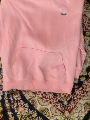 Rosa Lacoste hoodie - Säljer en snygg rosa hoodie från Lacoste i regular fit. Hoodien har en klassisk känguruficka framtill och den ikoniska gröna krokodilloggan på bröstet. Tillverkad i mjukt bomullsmaterial som känns skönt mot huden. Perfekt för dig som gillar stilrena och bekväma plagg.