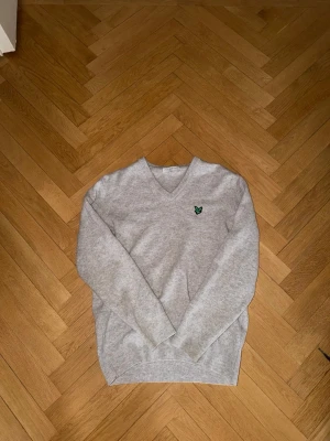 Grå v-ringad tröja från Lyle & Scott - Snygg grå v-ringad tröja från Lyle & Scott med deras klassiska gröna fågel-logga broderad på bröstet. Tröjan har långa ärmar och är tillverkad i mjukt stickat material som passar perfekt till lager-på-lager-stil.