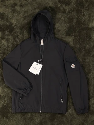 Svart Moncler vindjacka - Svart vindjacka från Moncler med huva och dragkedja framtill. Jackan har två fickor med dragkedja, elastiska muddar och Moncler-logga på ärmen. Tillverkad i lätt och slitstarkt material, perfekt för blåsiga dagar. Skickar samma dag rekommenderar PostNord går oftast snabbast. Skriv om du har frågor svarar så fort jag kan. Nfc tag funkar👍
