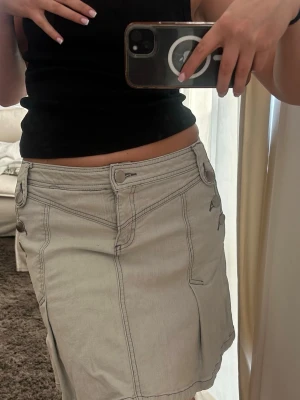 Ljus grå/beige kort jeanskjol med detaljer - Säljer en ljus grå/beige kort jeanskjol med coola sömdetaljer och fickor fram. Kjolen har knapp- och dragkedjestängning samt bälteshällor. Perfekt för dig som gillar en avslappnad men trendig stil.