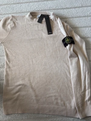Beige stickad tröja Stone Island - Snygg beige stickad tröja från Stone Island med klassisk rund hals och ribbade muddar. Tröjan har den ikoniska Stone Island-loggan på ärmen och är tillverkad i mjukt material som passar perfekt till alla outfits. Står XL men passar som S-M