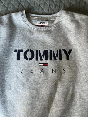 Grå sweatshirt från Tommy Jeans - Snygg grå sweatshirt från Tommy Jeans med stor logga framtill i svart, vitt och rött. Klassisk rund halsringning och mjukt material på insidan. Perfekt för en avslappnad och trendig look.
