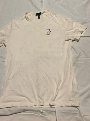 Vit t-shirt med Snoopy från H&M - Vit t-shirt från H&M med en liten Snoopy-brodyr på bröstet. Klassisk rund hals och korta ärmar. Tillverkad i mjuk bomull för en bekväm känsla. Perfekt för dig som gillar stilrena plagg med en rolig detalj.