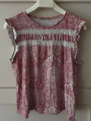 Rödvit ärmlös blus med coolt mönster  - Säljer en ärmlös blus med rödvitt blommigt mönster och spetsdetaljer framtill. Toppen har volangkant vid axlarna och rund halsringning. Perfekt för dig som gillar romantiska detaljer och boho-vibe. Står att det är en L men passar mer som S/M