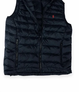 Svart dunväst från Ralph Lauren - Snygg svart dunväst från Ralph Lauren med quiltad design och dragkedja framtill. Västen har två praktiska fickor med dragkedja och den klassiska röda loggan broderad på bröstet. Perfekt för lager-på-lager och streetstyle. Den har aldrig använts och är helt ny skriv för mer info