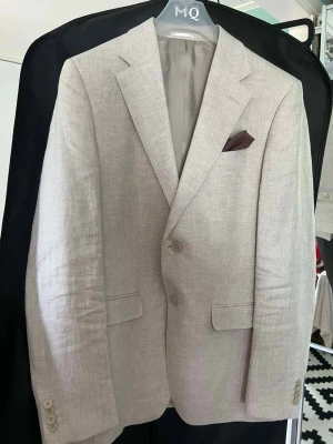 Beige linnekostym från MQ - Snygg beige kostym från MQ i linne. Kavajen har två knappar, klassiska slag och en bröstficka med mörkbrunt näsduk. Byxorna har raka ben och passpoalfickor bak. Perfekt för dig som vill ha en stilren och luftig look.