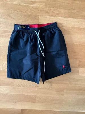 Svarta badshorts från Polo Ralph Lauren -   Polo Ralph Lauren med röd logga broderad på benet. De har elastisk midja med vit dragsko och röd insida i midjan. Praktiska fickor på sidorna och en bakficka med lock. Perfekta för stranden eller poolen.