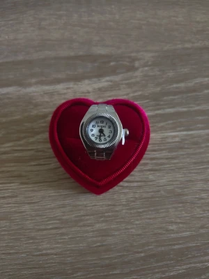 Silverfärgad ring med klocka - Unik silverfärgad ring med inbyggd klocka. Ringen har en rund urtavla med tydliga svarta siffror och visare, samt en metallisk, räfflad kant. Perfekt accessoar för dig som vill sticka ut och kombinera stil med funktion.