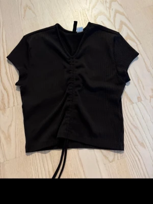 Svart ribbad croppad topp med snörning - Trendig svart ribbad topp från Monki med korta ärmar och croppad passform. Toppen har en snygg v-ringning och snörning framtill som ger en cool detalj. Perfekt att styla med höga jeans eller kjol.