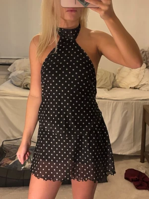 Svart prickig halterneckklänning - Säljer en svart kort halterneckklänning med vita prickar och öppen rygg. Klänningen har en knytning i nacken och volangkant nedtill. Har även inbyggda shorts under 
