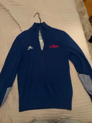 Blå halvzip golftröja från J.Lindeberg - Snygg blå golftröja med halv dragkedja från J.Lindeberg. USA-tryck på bröstet och ryggen, samt vita detaljer och ränder på ärmsluten och insidan av dragkedjan. Perfekt för golfen eller som ett sportigt vardagsplagg. Den är stickad och gjord av merinoull.