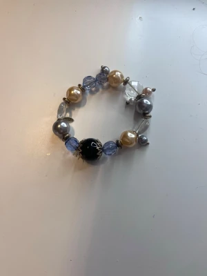 Armband med mixade pärlor - Unikt armband med pärlor i olika färger och former. Pärlorna är i nyanser av blått, svart, silver, vitt och guld, både matta och glansiga. Armbandet har elastisk tråd och blandade pärlor i plast och glas för en cool och personlig stil.