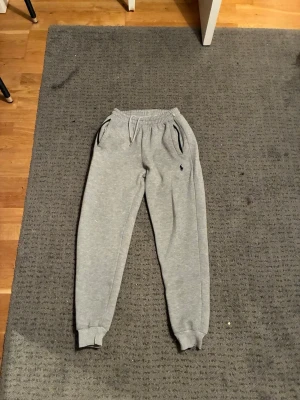 Grå mjukisbyxor från Polo Ralph Lauren - Säljer ett par klassiska grå mjukisbyxor från Polo Ralph Lauren med dragsko i midjan, muddar vid bensluten och två fickor med svarta detaljer. Liten broderad logga på vänster ben. Perfekta för chill dagar eller när du vill vara bekväm.
