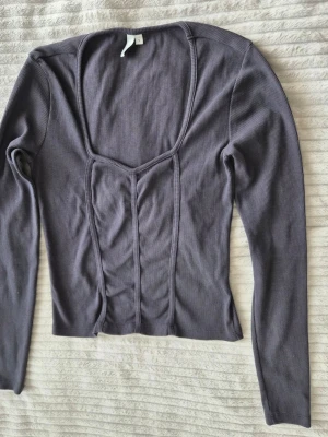 Lila ribbad långärmad topp - Trendig lila ribbad topp med lång ärm och markerade sömmar framtill som ger en korsettliknande look. Djup fyrkantig ringning och figurnära passform, perfekt att styla med jeans eller kjol.