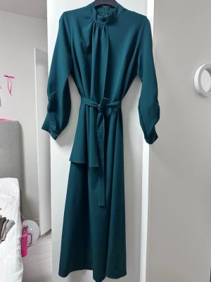 Mörkgrön långklänning med knytband - Snygg mörkgrön långklänning med markerad midja och knytband. Klänningen har långa ärmar med lätt puff och resår vid ärmslut samt hög halsringning. Klänningen ser blå ut i bilden, men är mörkgrön.