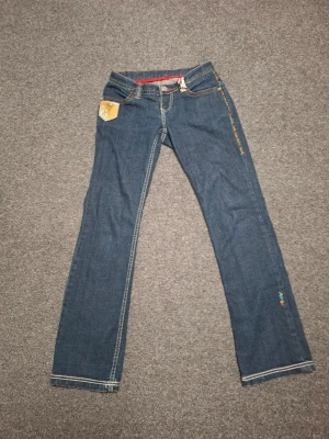 Blå jeans med broderier och patch - Snygga blå jeans med raka ben, dekorativa broderier längs ena benet och en gul patch på fickan. Jeansen har kontrastsömmar och en röd detalj i midjan. Perfekta för dig som gillar unika detaljer och streetstyle.